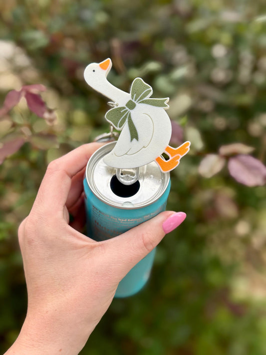 May include: A white goose-shaped can opener with an orange beak and feet, and a green bow. The opener is attached to a silver keychain and is placed on top of a blue can. The background is blurred green and pink foliage.
