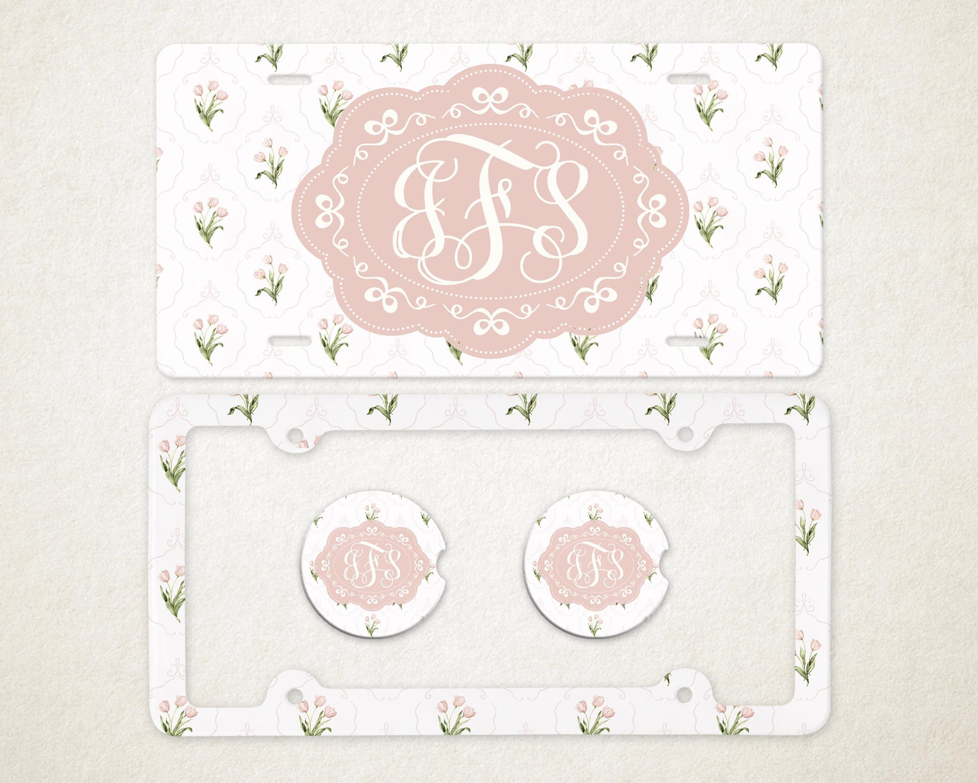 May include: A license plate, license plate frame, and two car coasters with a floral pattern and a monogram. The license plate and coasters have a pink oval with a white monogram. The frame and plate have a white background with pink flowers.