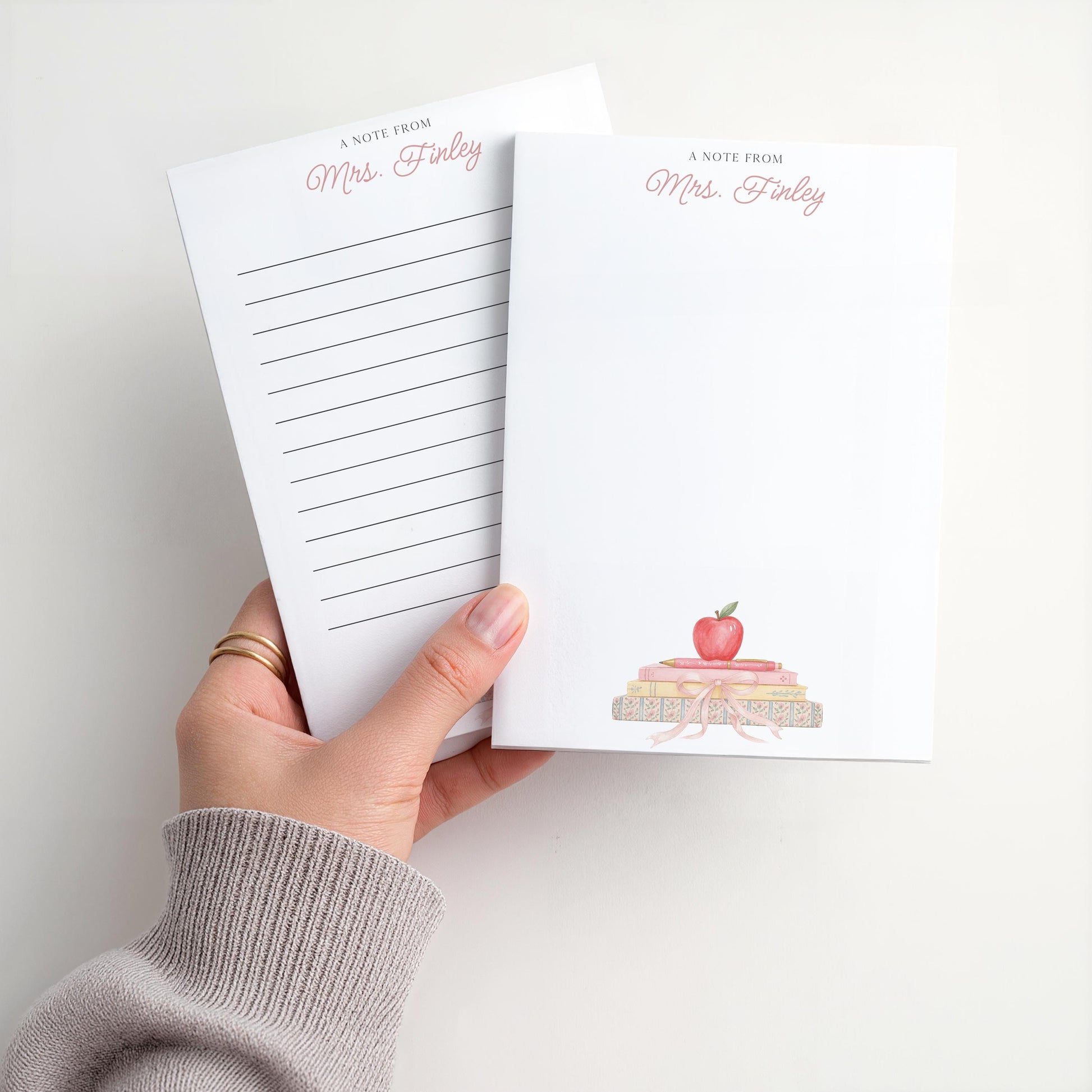 May include: White notepad with the text "A Note From Mrs. Finley" in pink script. The notepad features a watercolor illustration of a stack of books topped with a red apple. One notepad has lined pages.